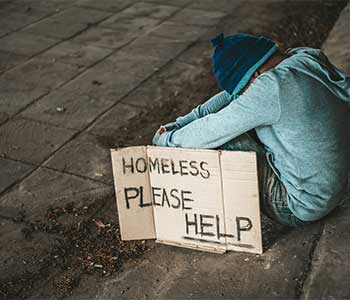 A person sitting on the ground with a cardboard sign that reads "HOMELESS PLEASE HELP.